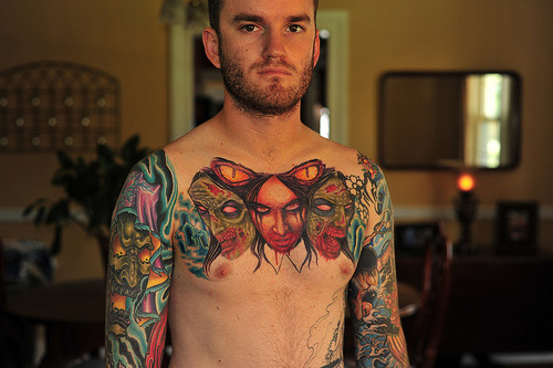 A shirtless man with large colorful tattoos across his chest and down his 